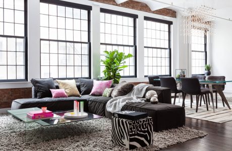 Spacious loft-style living room with large windows, black sofas, and modern chandelier.