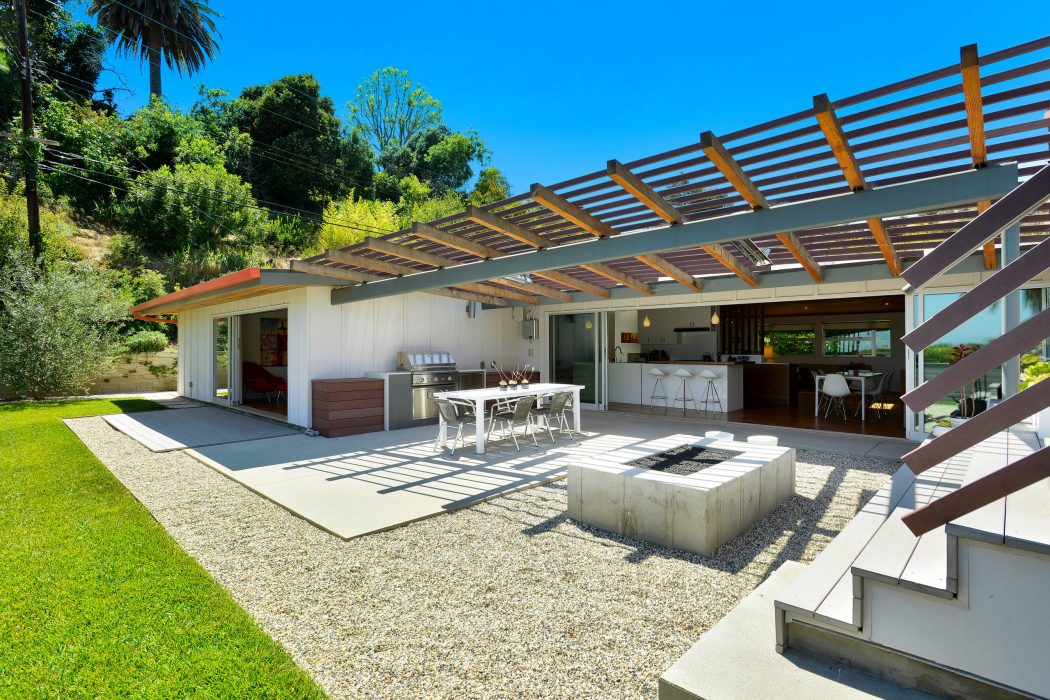 A modern outdoor living space with a covered patio, kitchen, and dining area surrounded by lush greenery.