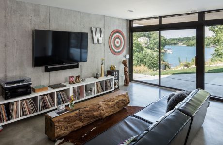 Modern living room with concrete walls, wood accents, and floor-to-ceiling windows overlooking a lake.