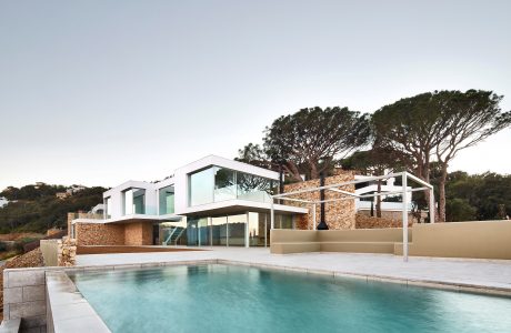 Modern villa with sleek glass walls, stone exterior, and an infinity pool surrounded by lush greenery.