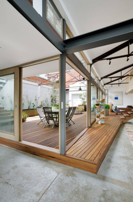 Contemporary indoor-outdoor living space with wooden deck, glass walls, and industrial overhead beams.