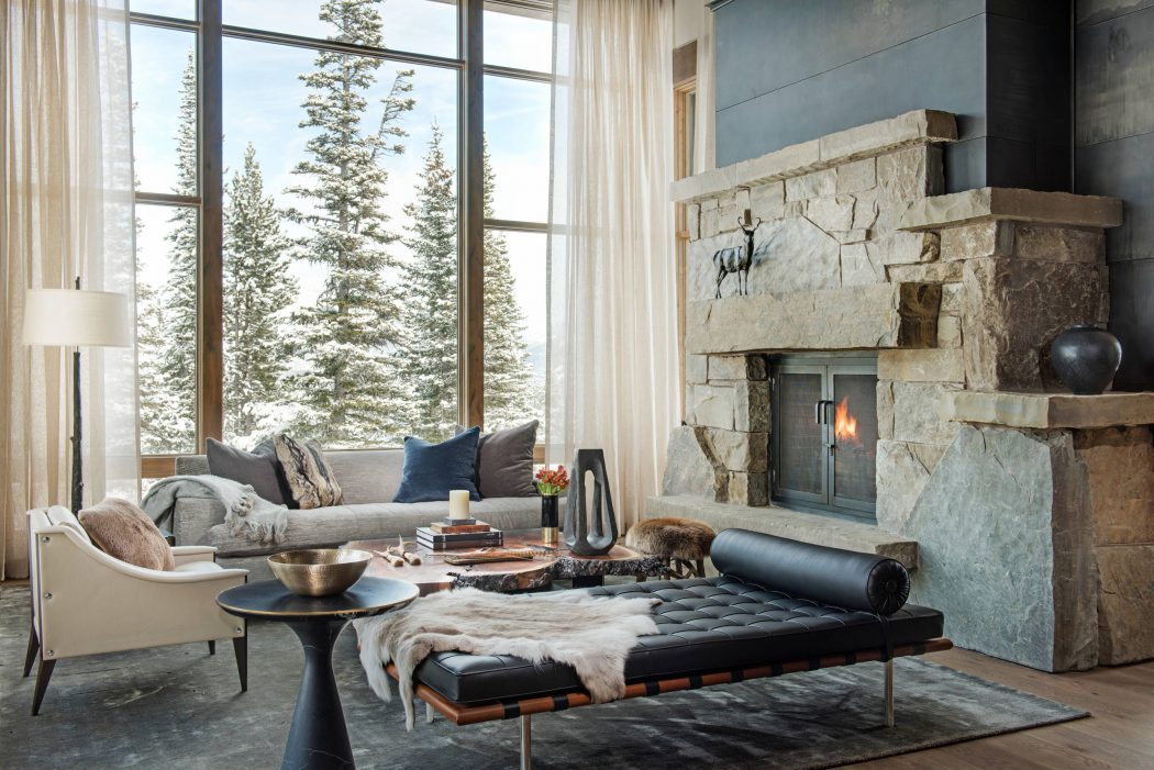 Cozy, rustic living room with stone fireplace, large windows overlooking snowy trees.