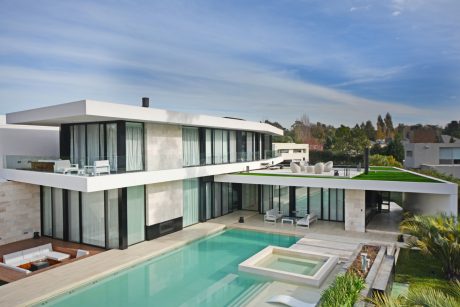 Modern, minimalist home with glass walls, elevated pool, and lush landscaping.