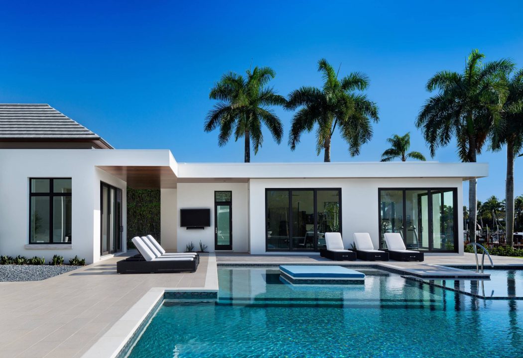 A modern, open-concept home with a pool, sun loungers, and lush palm trees.