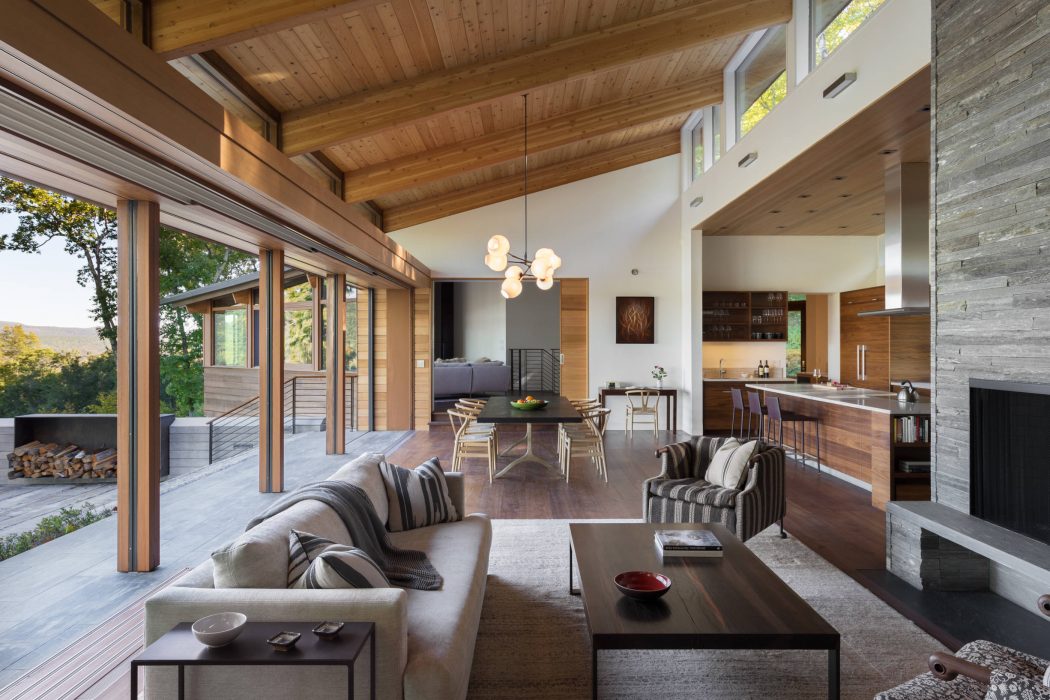 Contemporary living room with high ceilings, wooden beams, and floor-to-ceiling windows overlooking nature.