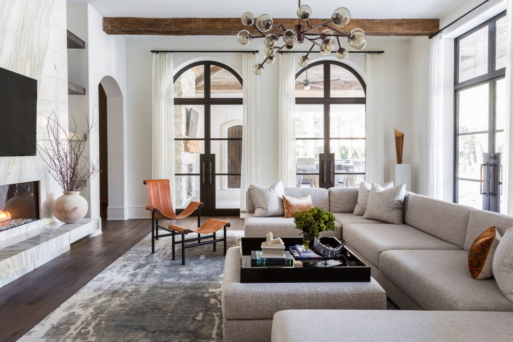 A cozy living room with arched windows, a rustic beam ceiling, and a contemporary chandelier.