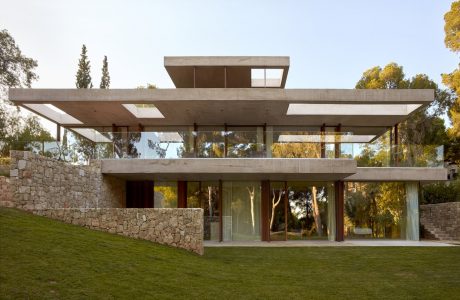 A modern concrete and glass structure with a cantilevered roof and stone walls, surrounded by lush greenery.