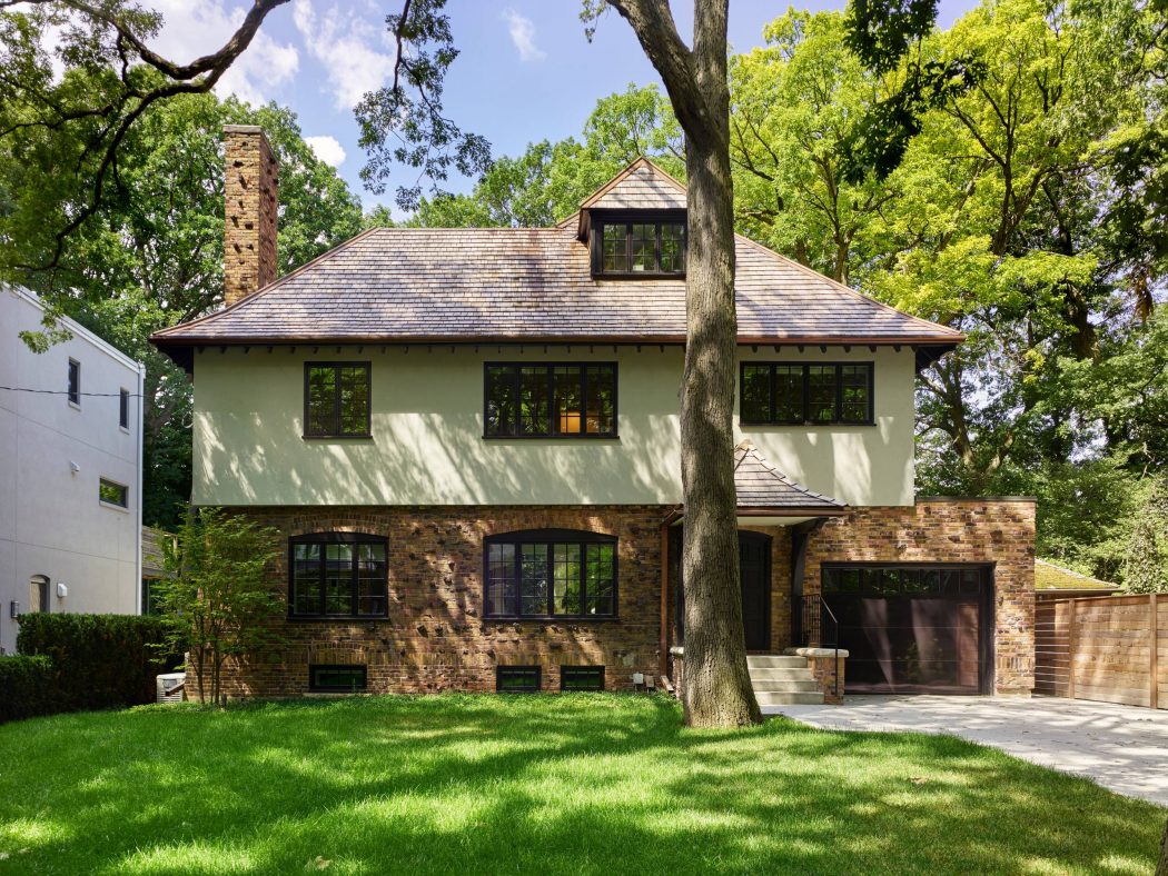 A two-story house with a stone exterior, slanted roof, and large windows surrounded by lush greenery.
