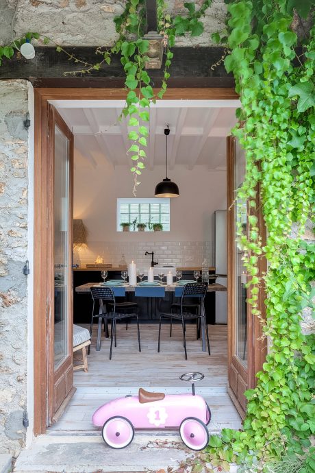 A rustic stone building with a dining area, greenery, and a pink toy car on the porch.