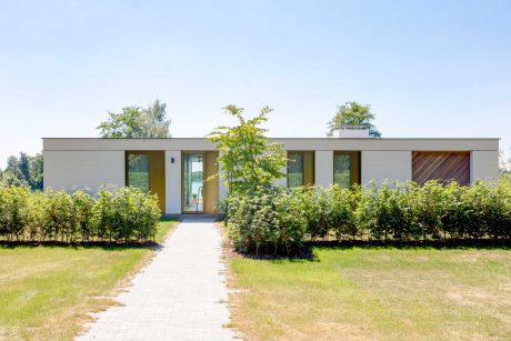 Minimalist, single-story home with large windows and a paved walkway surrounded by lush greenery.
