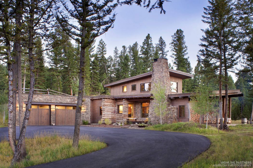 Rustic mountain lodge with stone chimney, wooden siding, and wraparound porch in forested setting.