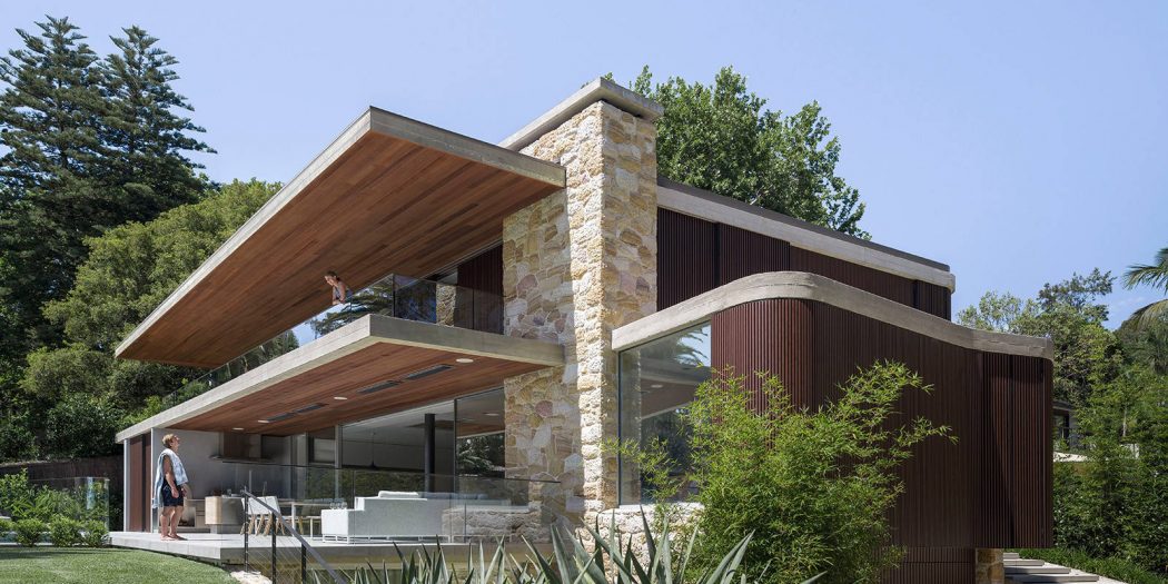 A modern, wood-and-stone home with large windows, overhanging roof, and lush greenery.