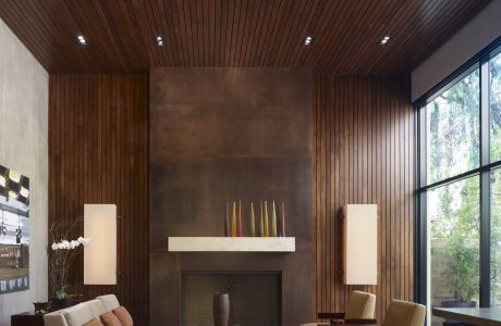 Warm, modern living room with wood-paneled ceiling, concrete fireplace, and large windows.