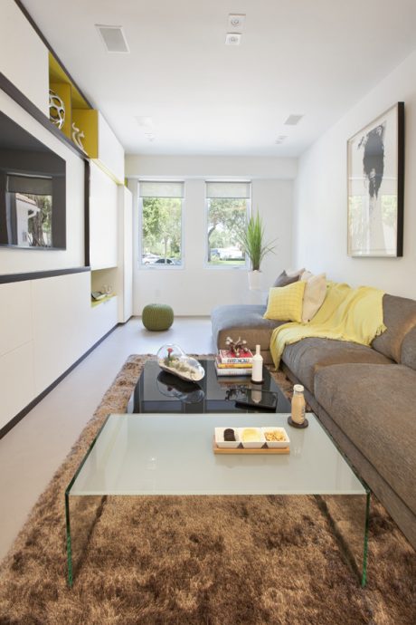 Modern living room with white walls, gray sofa, yellow accents, and glass coffee table on a shag rug.