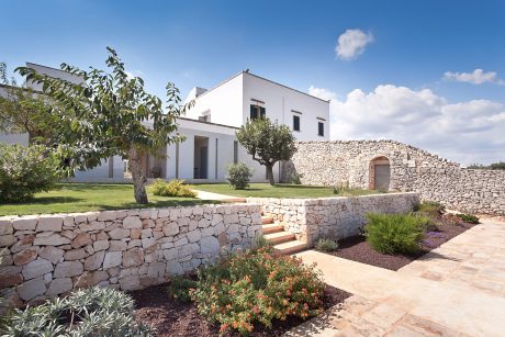 A modern Mediterranean-style villa with a stone facade, surrounded by a lush garden and landscaping.