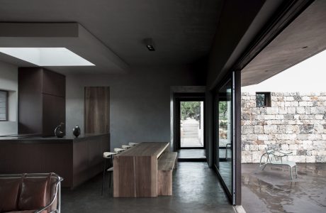 A sleek and modern interior with a wooden kitchen counter, concrete flooring, and large windows overlooking a stone wall and outdoor path.