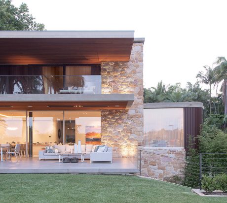 A modern, open-concept home with a stone facade, wooden beams, and lush greenery.