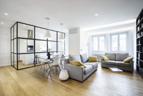 Modern open-concept living room and kitchen with sleek black-framed glass walls and minimalist furnishings.