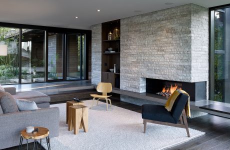 Contemporary living room with stone fireplace, built-in shelves, and large glass windows overlooking nature.