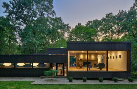 Modern cabin-style home with clean lines, large windows, and a wooden deck surrounded by lush greenery.