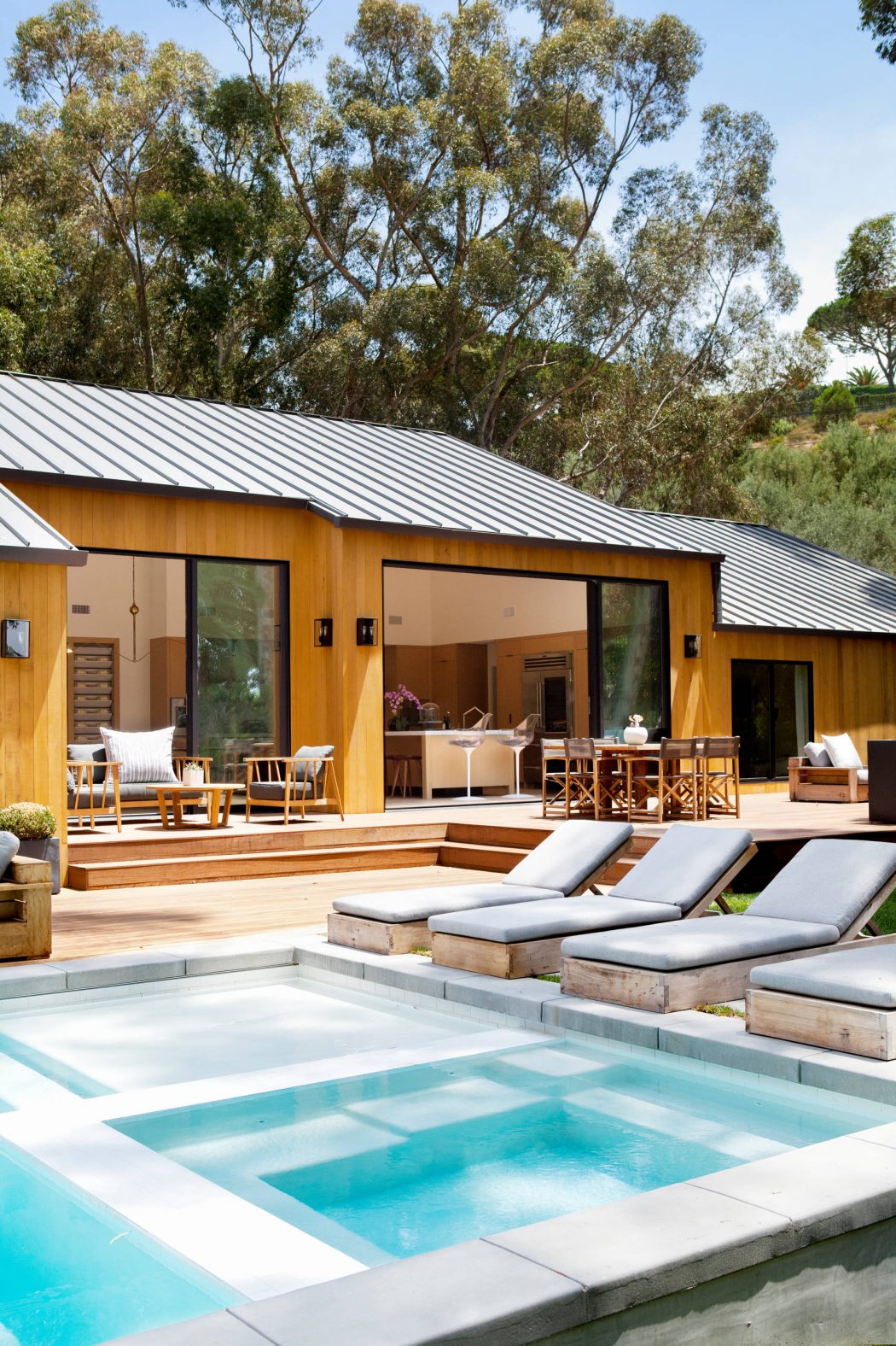 Modern wooden cabin with a sleek metal roof, surrounded by lush greenery and a tranquil pool.