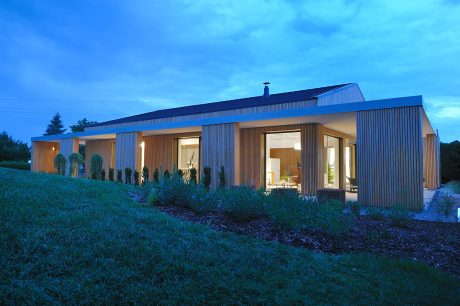 Modern wooden-clad structure with large glass windows overlooking a grassy slope at dusk.