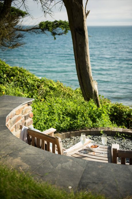 A cozy outdoor seating area overlooking the ocean, with a stone fireplace and wooden furniture.