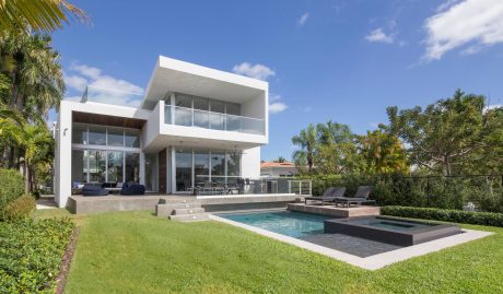 Modern, two-story house with glass walls, pool, and lush tropical landscaping.