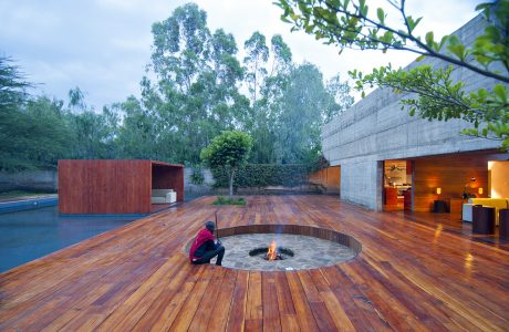 Sleek, minimalist outdoor space with a circular firepit on a wooden deck, surrounded by lush greenery.