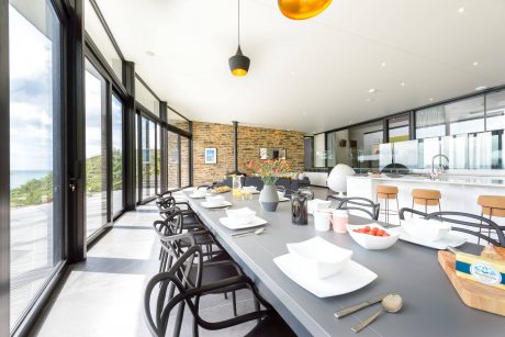 Modern open-plan kitchen and dining area with floor-to-ceiling windows, stone walls, and pendant lights.