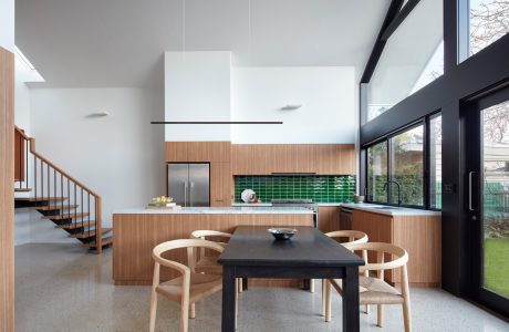 Modern open-plan kitchen with natural wood cabinetry, green tile backsplash, and large dining table.