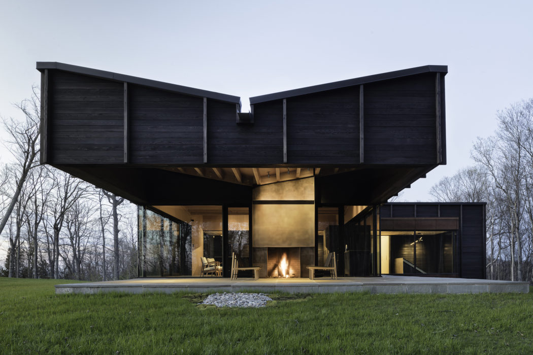 A modern, angular cabin with a prominent central fireplace and expansive windows facing a forested landscape.