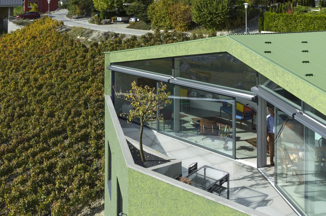 A modern, glass-enclosed structure with a green, sloping roof overlooks a lush landscape.