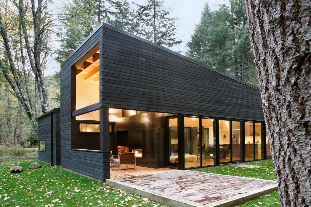 A modern black cabin nestled in a lush forest, featuring large glass windows and a wooden deck.