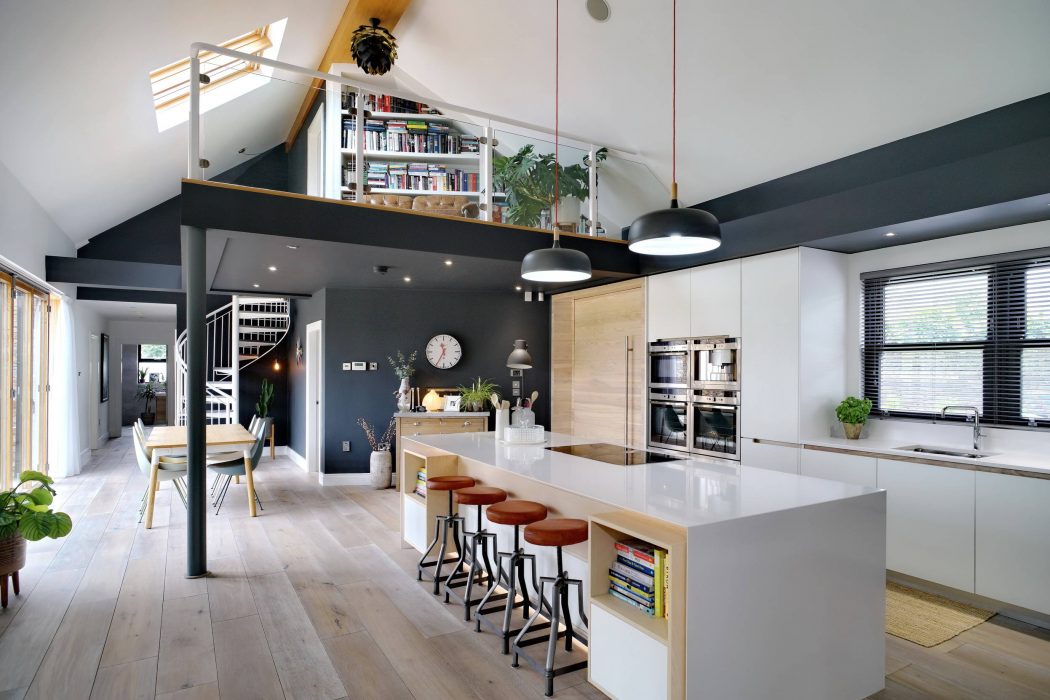 A modern, open-plan kitchen with a sleek white island, exposed beams, and a mezzanine level.