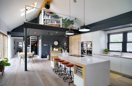 A modern, open-plan kitchen with a sleek white island, exposed beams, and a mezzanine level.