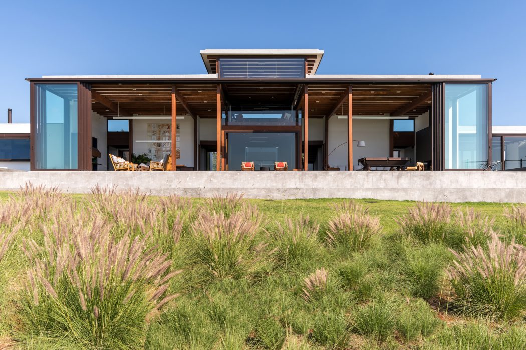 A modern, open-concept home with a large covered patio, wood and glass facade, and lush grass landscaping.