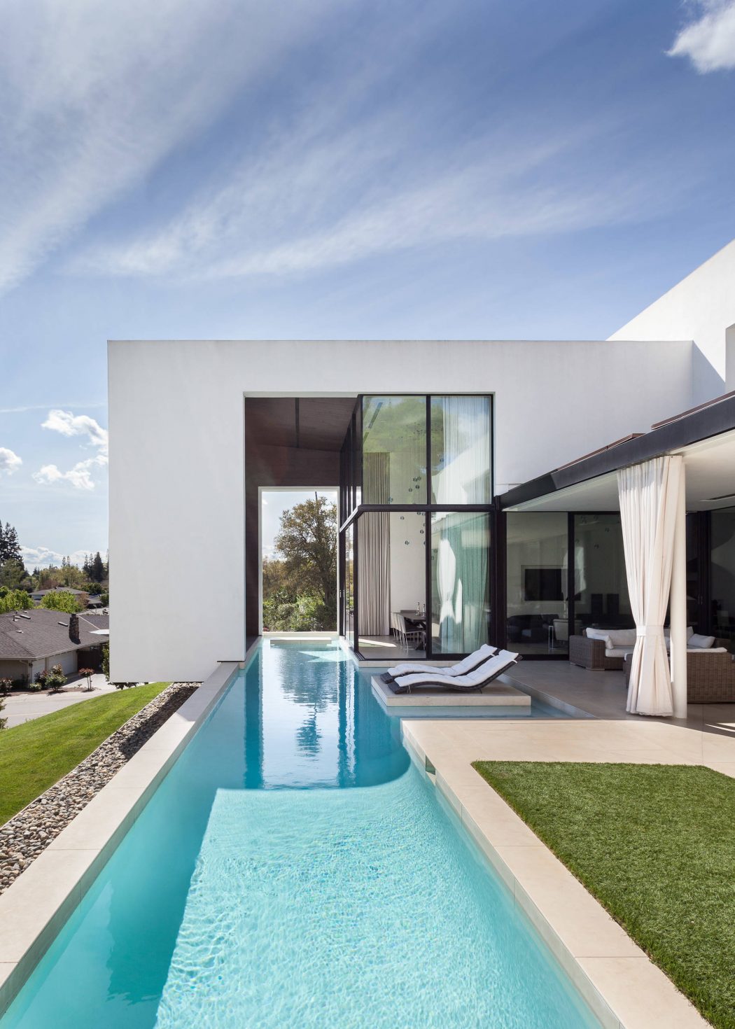 Sleek, modern home with a long, narrow pool, surrounded by lush greenery and clear skies.