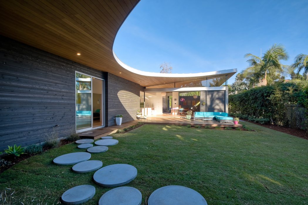 Curved wooden exterior with floor-to-ceiling glass, lush landscaping, and stepping stones.