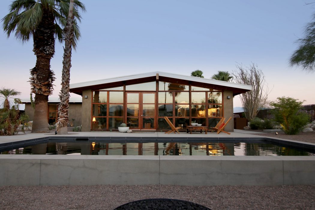 Modern desert home with glass walls, pool, and outdoor seating area against palm trees.