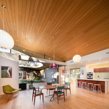 Warm, modern interior with sleek wood paneled ceiling, minimalist furniture, and floor-to-ceiling windows overlooking lush greenery.