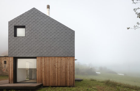 A modern, wood-clad house with a gray shingled roofline stands in a foggy, grassy field.