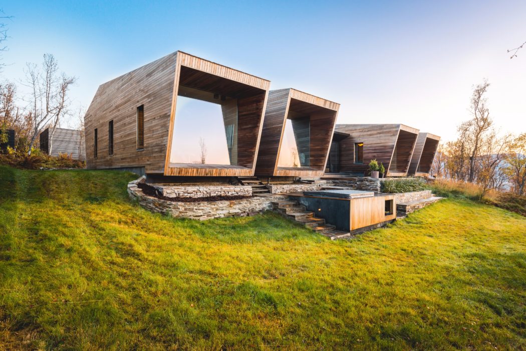A modern, wooden structure with asymmetrical windows and a stone foundation sits in a grassy field.