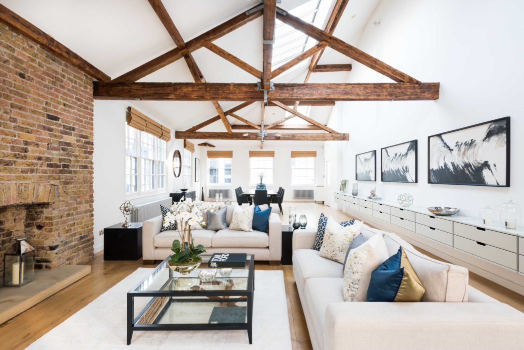 A modern, open-concept living space with exposed wooden beams, brick walls, and large windows.