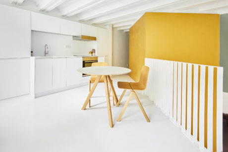Clean, minimalist kitchen with white cabinetry, wooden table, and vibrant yellow accent wall.