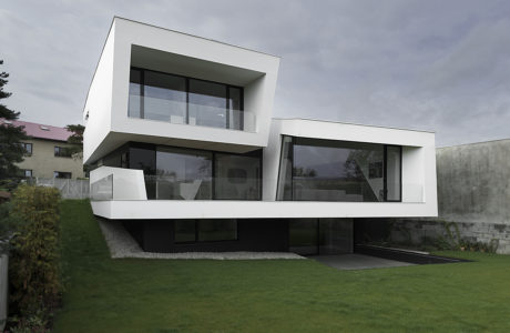 A modern, two-story residential building with a clean, minimalist exterior design featuring large glass windows and a cantilevered second floor.