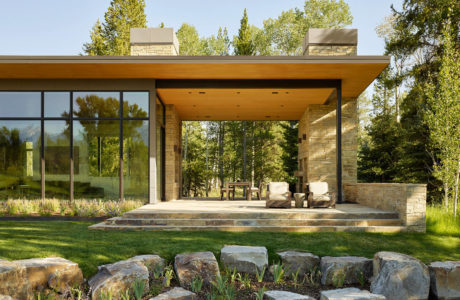 A modern, open-concept home with large glass windows, wood beams, and stone accents surrounded by lush greenery.