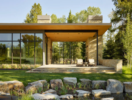 A modern, open-concept home with large glass windows, wood beams, and stone accents surrounded by lush greenery.