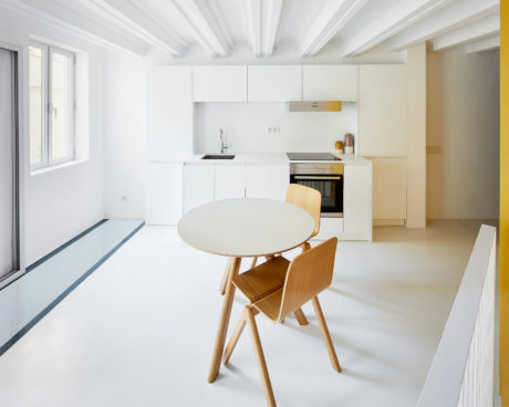 A modern, minimalist kitchen with clean white cabinets, wooden chairs, and a round dining table.