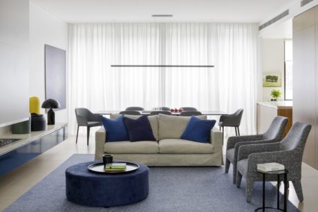 Modern, minimalist living room with neutral-toned furniture, floor-to-ceiling drapes, and a sleek linear lighting fixture.